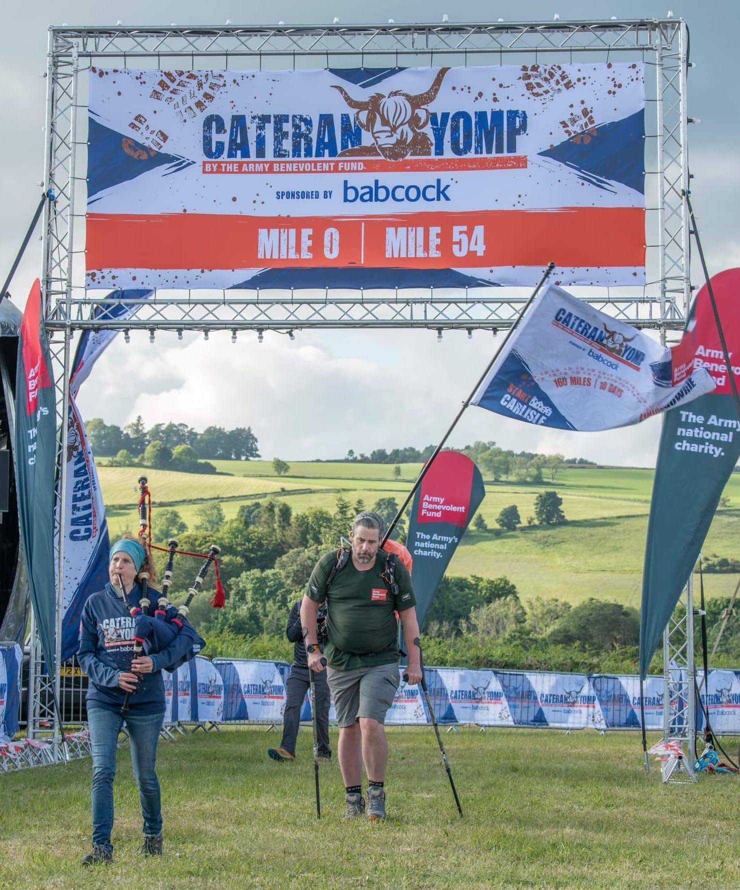 Veteran Mark Harding crossing Cateran Yomp finish line, beneath a banner and on crutches, accompanied by a bagpiper.