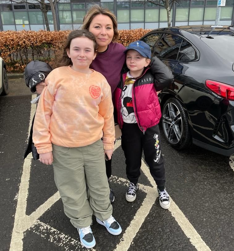 Mother with arms around daughter and son, pictured in car park