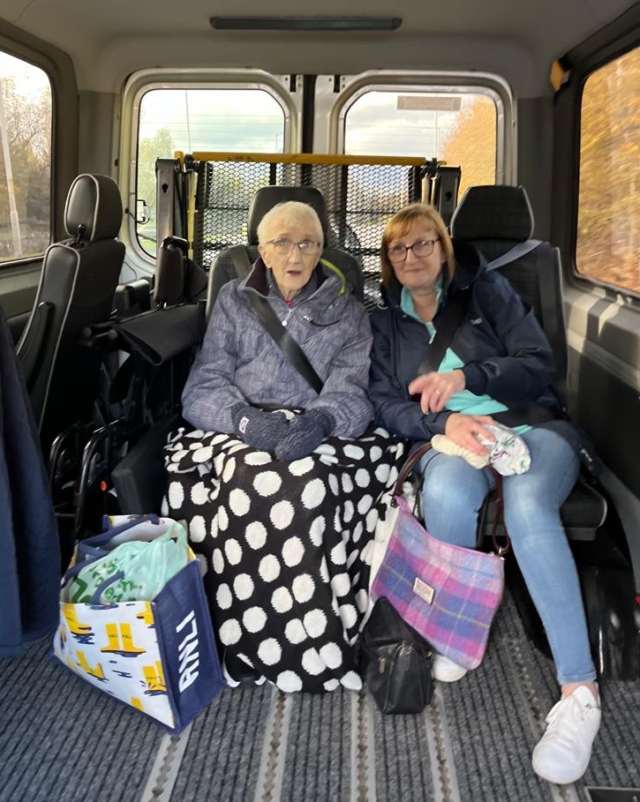 Elderly woman seated in wheelchair taxi, with female volunteer sitting next to her